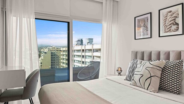 A modern hotel room with a large bed, a desk, and a chair. There are two framed pictures on the wall, and a balcony with a view of the city. -  Limassol Seaside Residences 06 (Limassol)