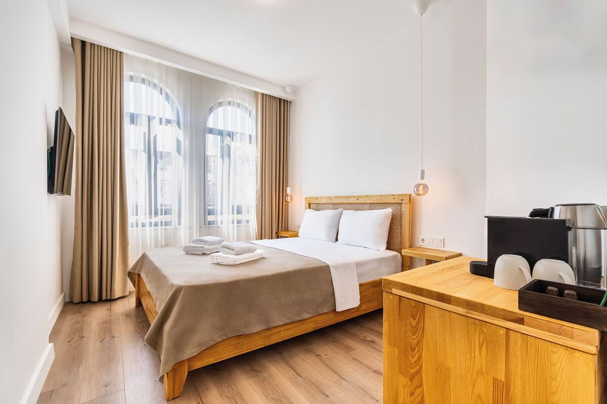 A hotel room with a bed, wooden bedside table, and a small kitchenette area.