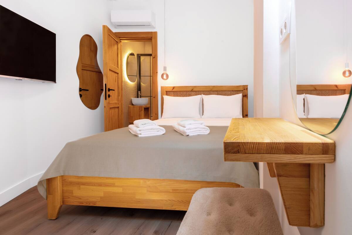 A modern hotel room with a wooden bed, a wooden desk, a flat-screen TV mounted on the wall, and a large mirror.