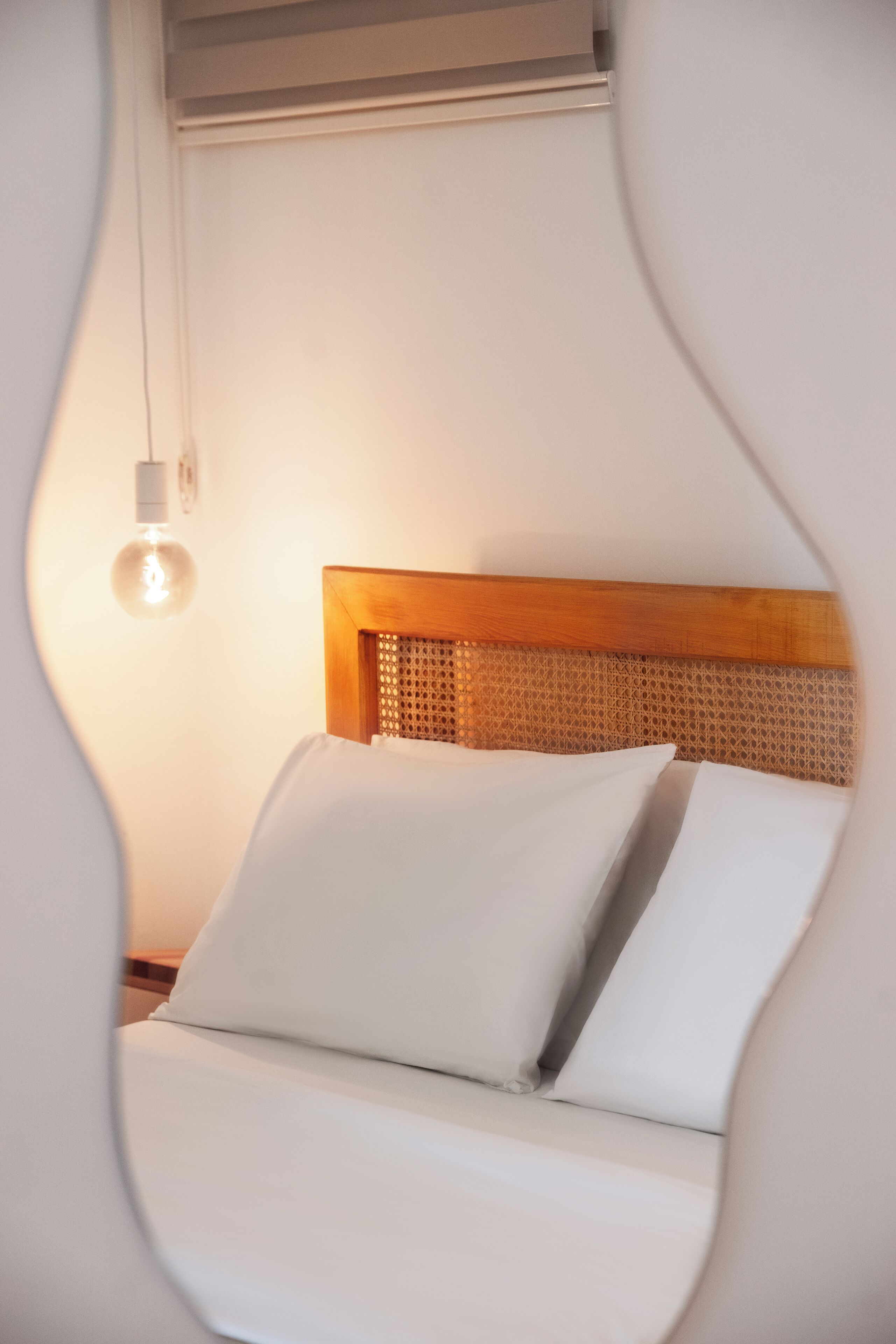 Een bed met witte lakens en een houten hoofdbord, een hangende lichtbol en een stopcontact aan de muur.