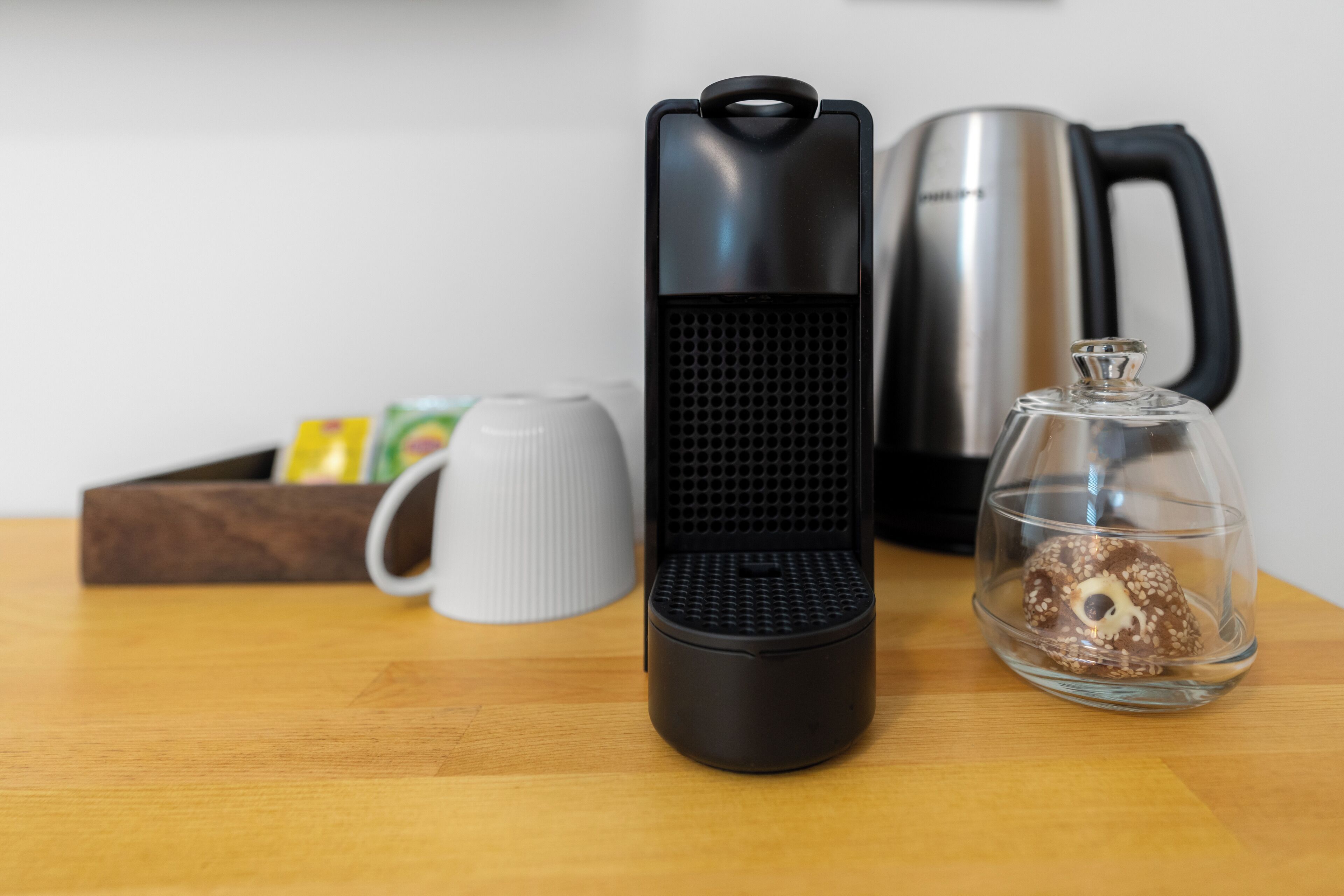 A kitchen area with a coffee maker, a kettle, a mug, and a glass container with a donut.