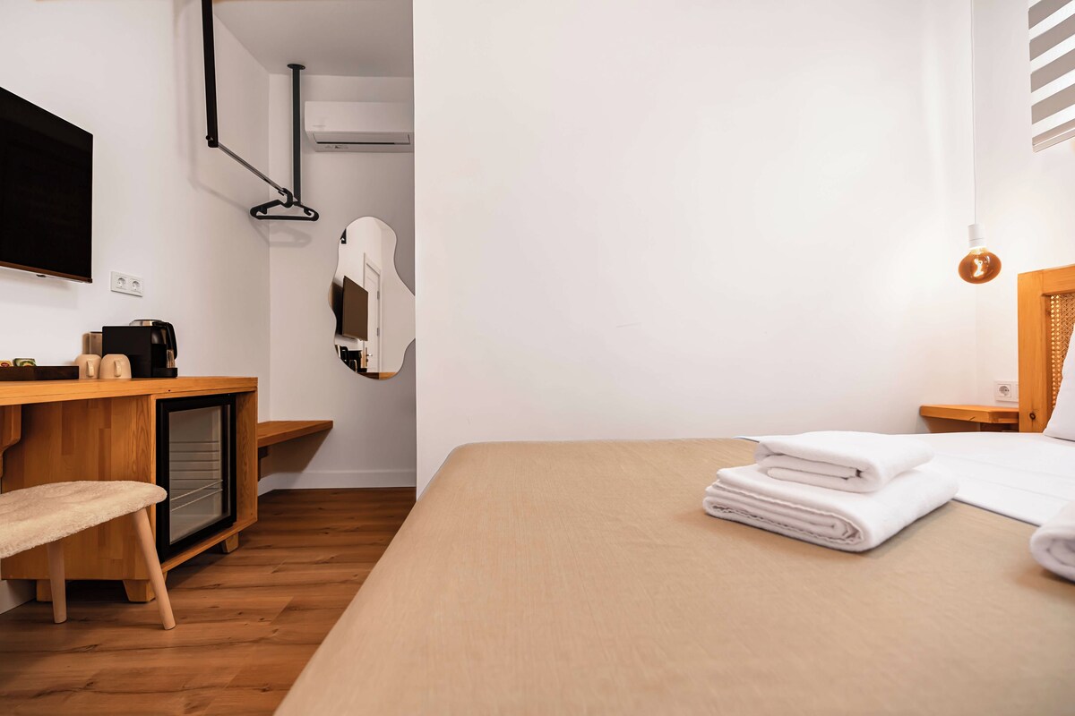 Habitación de hotel minimalista con cama, escritorio, silla, televisor y perchero.