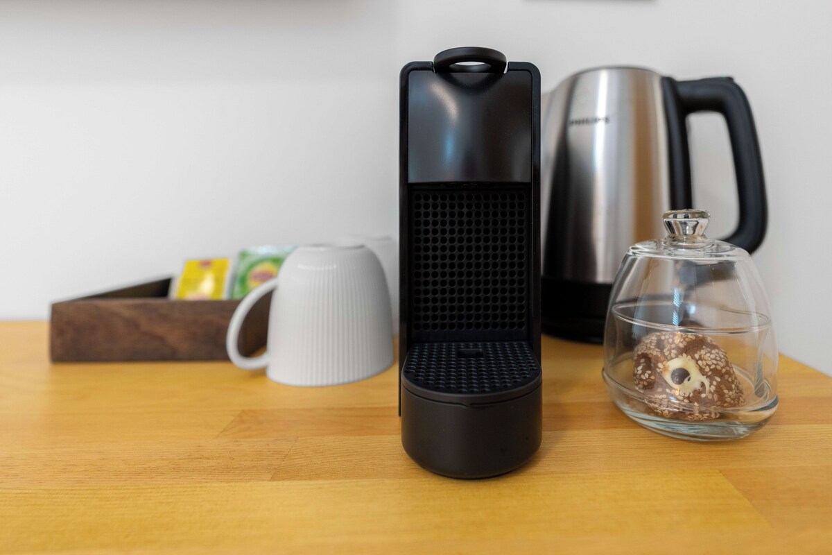 Una encimera de cocina con una cafetera, un hervidor, una taza, un frasco de vidrio con tapa y una caja de madera con objetos.