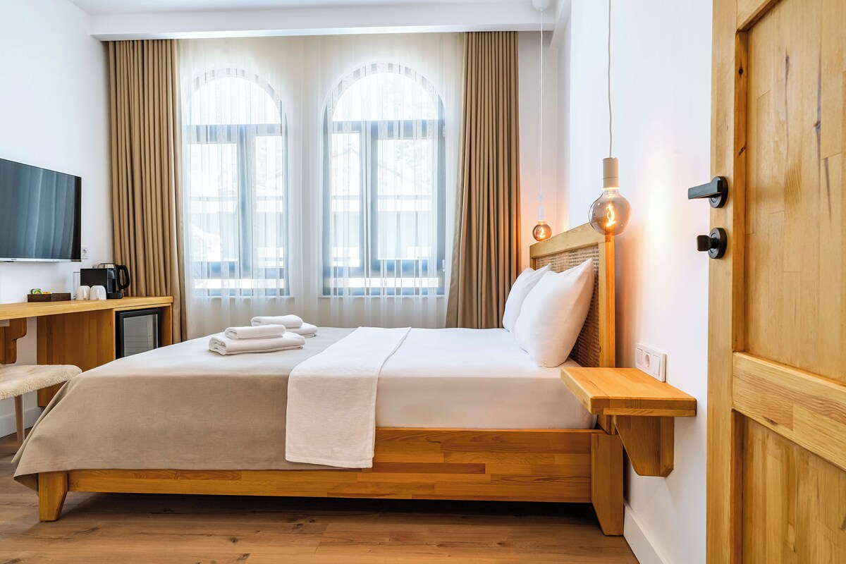 Habitación de hotel con cama de madera, un escritorio con televisor de pantalla plana, una mesita de noche con lámpara y ventanas grandes con cortinas.