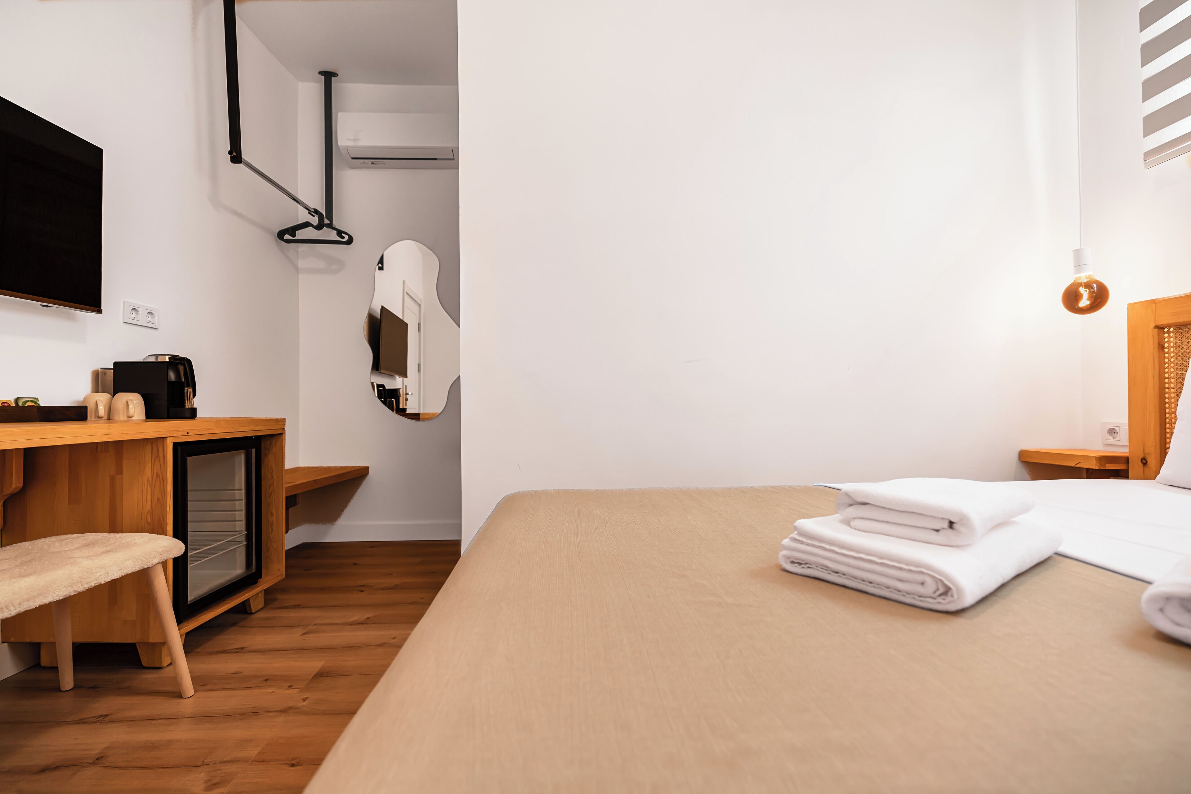 A modern hotel room with a wooden desk, a bed with white linens, a TV mounted on the wall, and a hanging coat rack.