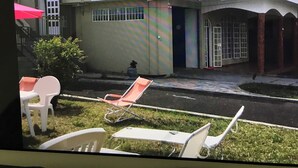 Un edificio de dos pisos con un balcón, un pequeño jardín con sillas y un cielo despejado.