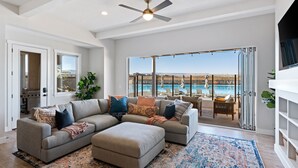 A spacious living room with a large sectional sofa, a ceiling fan, and a view of a pool through glass doors. - Persian Bliss: Desert Color Lagoon + Beaches (St. George)