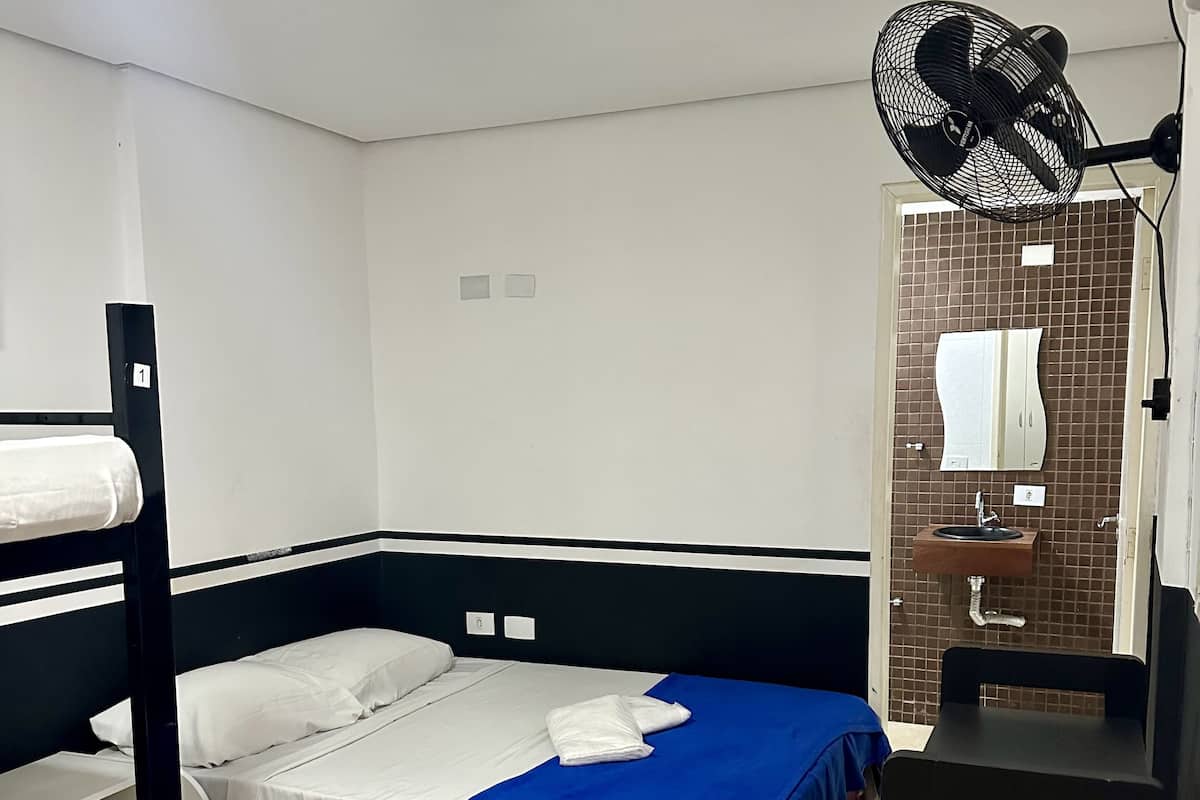 A single-bed room with a bunk bed, a desk, a chair, a fan, a mirror, and a bathroom with a tiled wall.