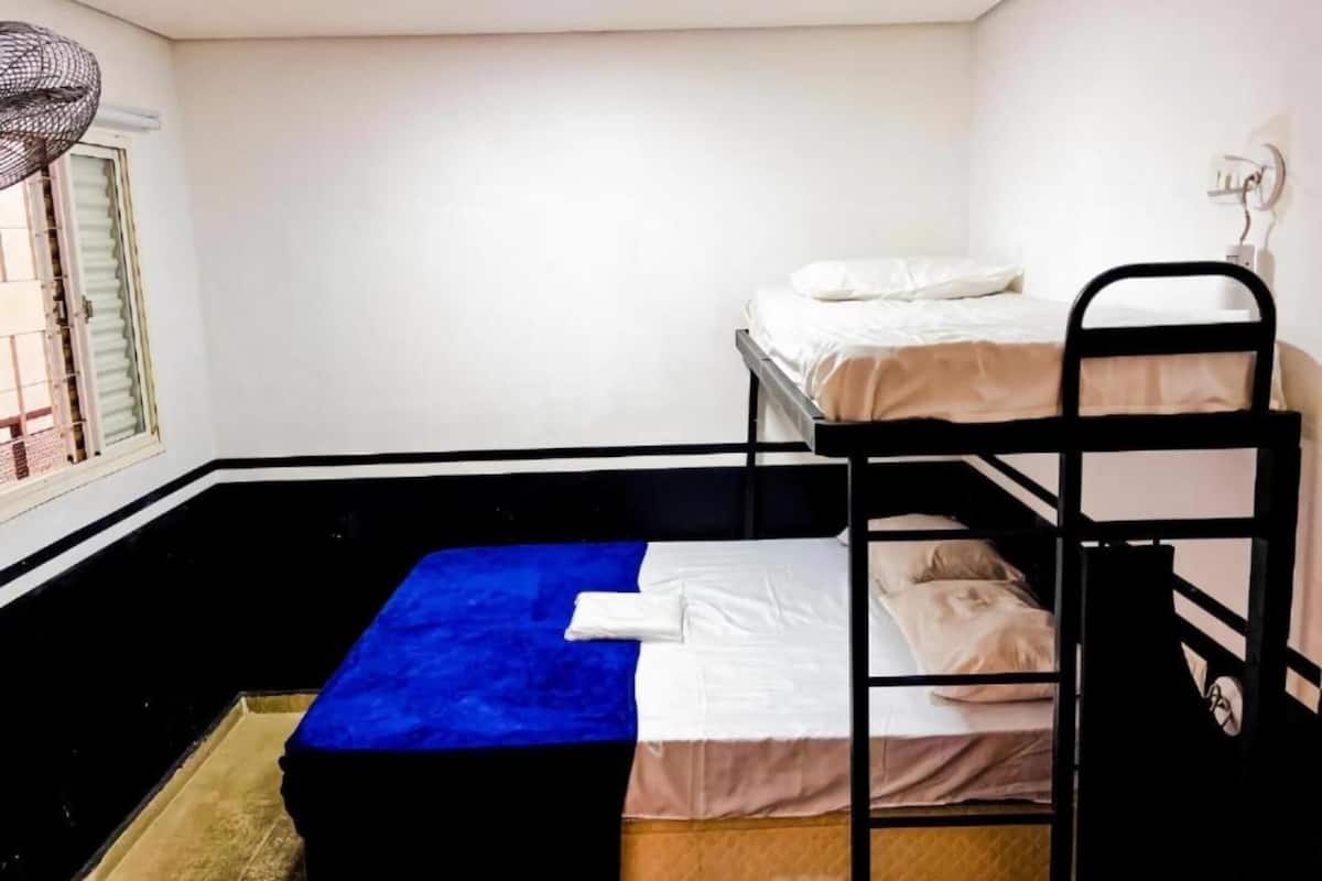 A dormitory room with bunk beds, a window with blinds, and a ceiling fan.