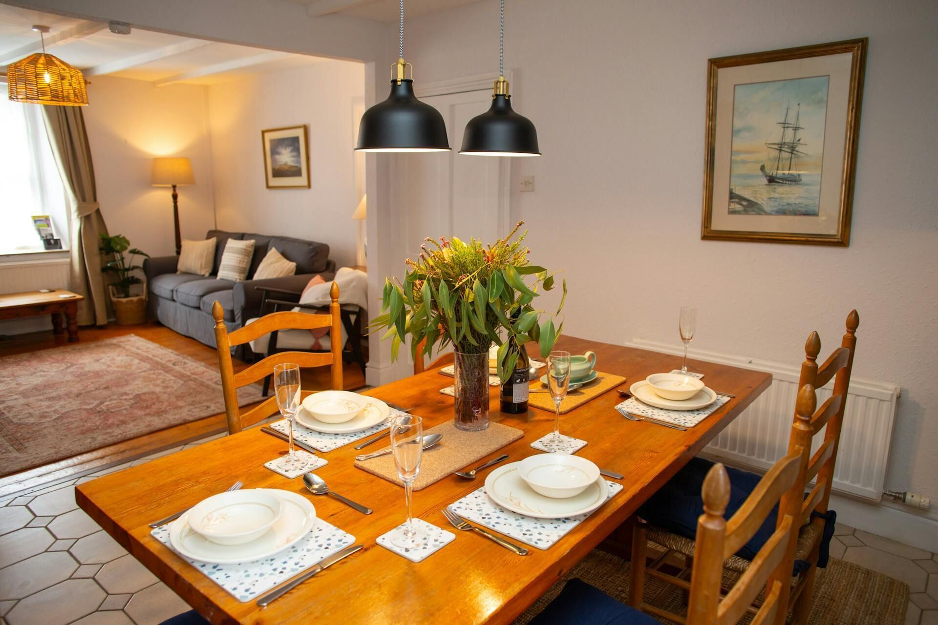 A dining area with a wooden table set for four, surrounded by wooden chairs. There is a vase with green foliage on the table, and a framed picture of a ship on the wall.