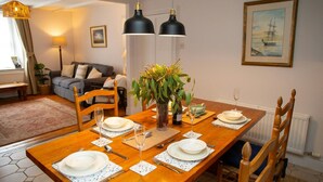 Een eethoek met een houten tafel voor vier personen, omringd door houten stoelen. Op de tafel staat een vaas met groene planten, en aan de muur hangt een lijst met een afbeelding van een schip.