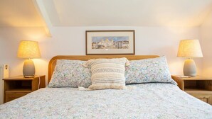 Een opgemaakt bed met bloemenbedrukking, twee bedlampjes, een lijst met een afbeelding aan de muur en een houten hoofdbord.