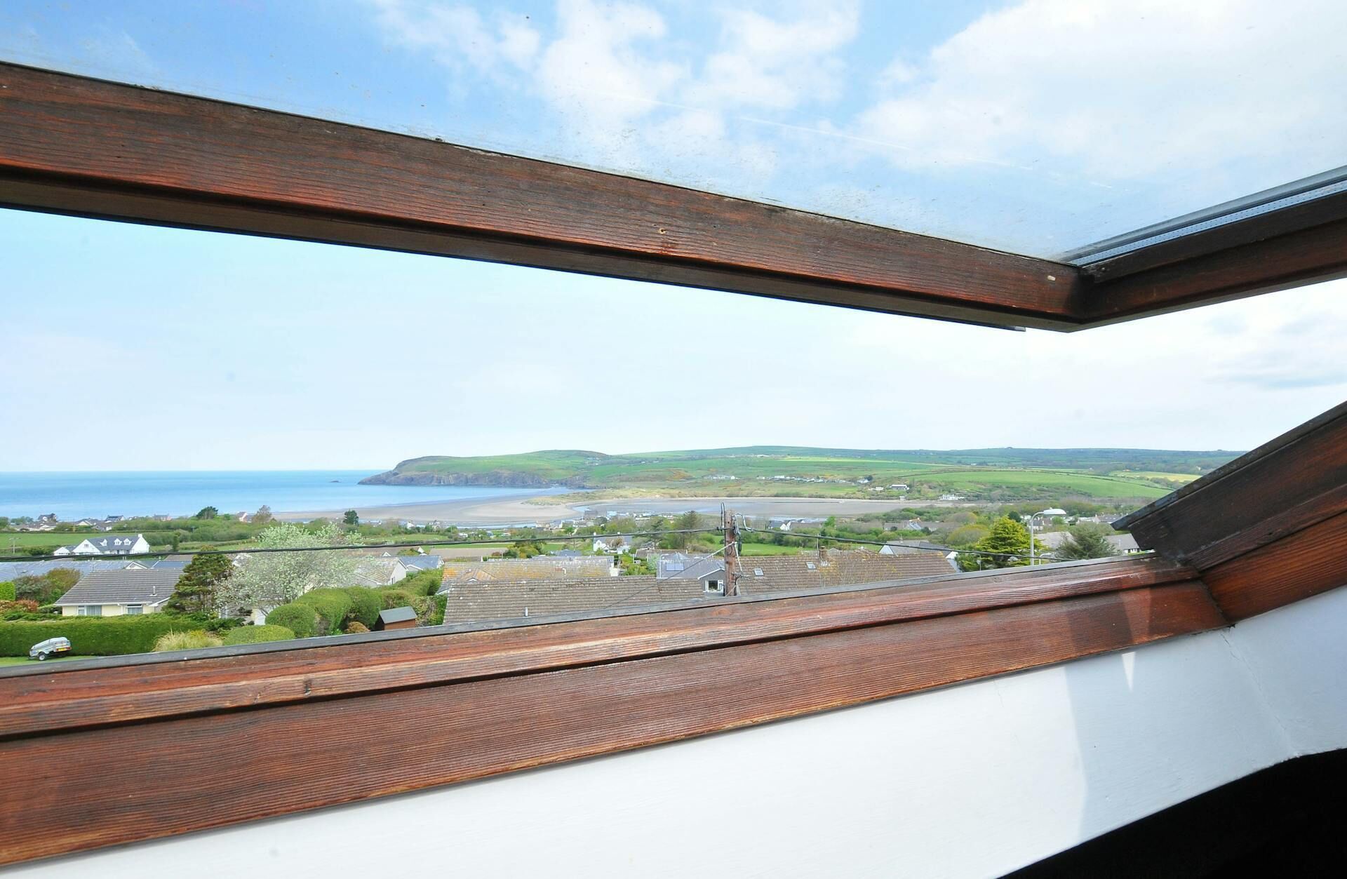 Vista desde una ventana con vigas de madera, que da a un paisaje costero con casas, vegetación y una playa.