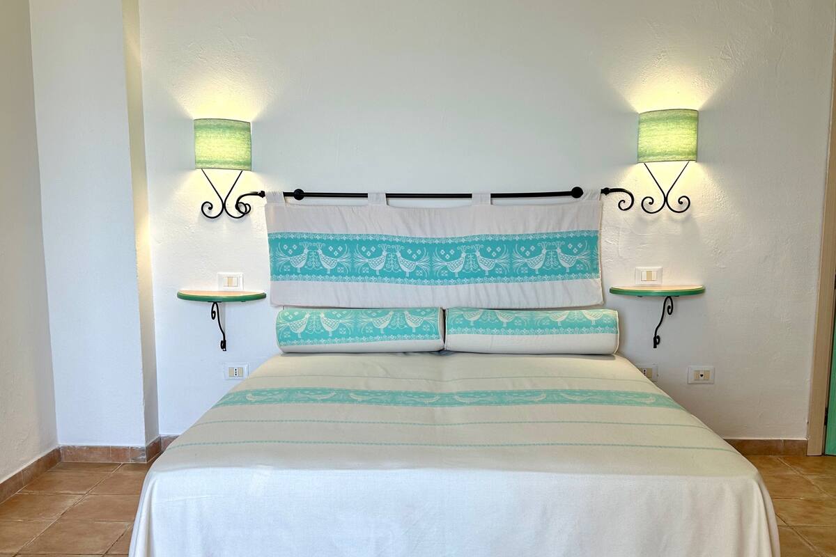 A neatly made bed with a white and teal striped bedspread, two matching pillows, and a headboard with a black horizontal bar. There are two bedside tables with lamps on either side of the bed.