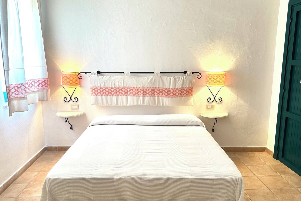 A hotel room with a bed, two wall-mounted lamps, a window with curtains, and a green door.