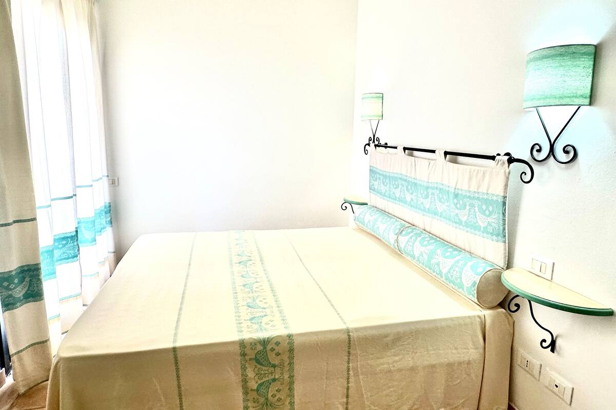 A bedroom with a bed, a bedside table, a wall-mounted shelf, and a window with curtains.