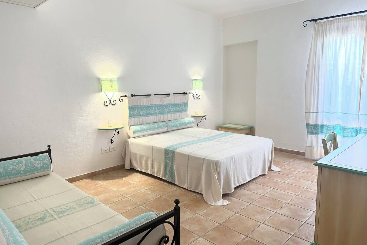 A bedroom with a bed, bedside tables, a nightstand, a window with curtains, and a tiled floor.