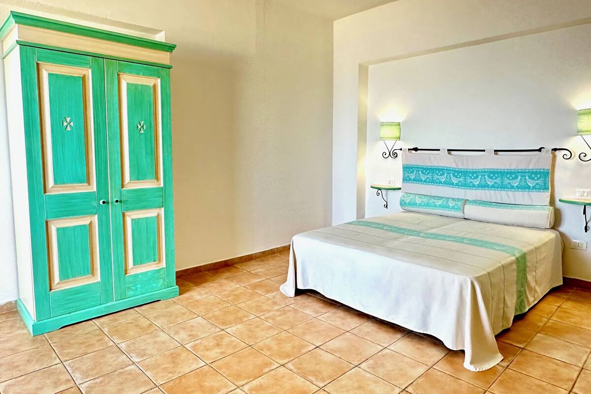 A bedroom with a bed, a green wardrobe, and a nightstand with a lamp.
