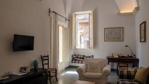 Un saló acollidor amb un sofà, un armari i una taula de fusta. Hi ha una televisió penjada a la paret, i una finestra amb vista a un balcó.