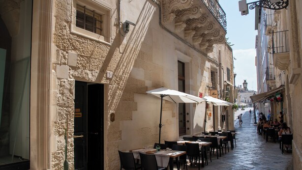 Un carrer empedrat estret amb terrasses de cafè, edificis de pedra i un cel clar.