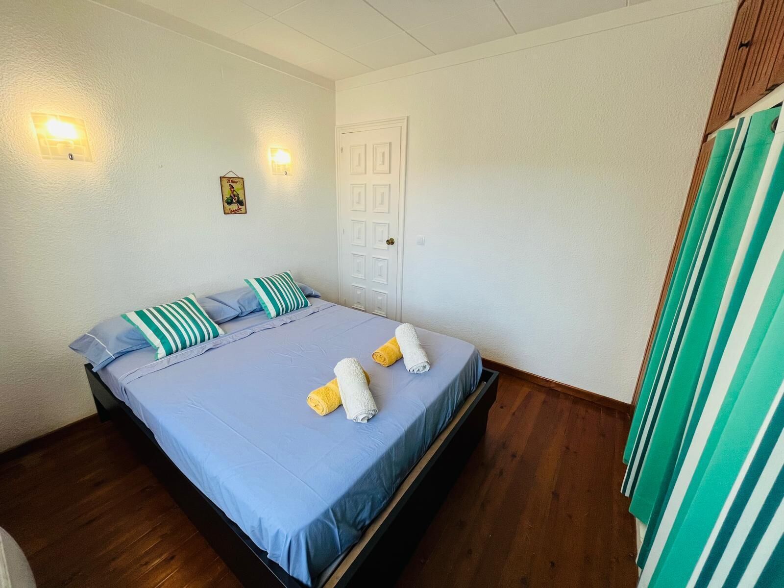 A single-bed room with a wooden floor, a white door, and a striped curtain.