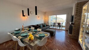 A modern dining area with a glass table, white chairs, and teal plates. A sofa and a balcony with a view are visible in the background. - Roses - Los Sauces - Apart 3 rooms (Rosas / Roses)