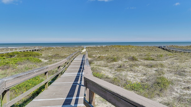 Een houten looppad dat naar een strand leidt, met duinen en een helderblauwe lucht.