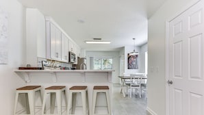 Een moderne keuken met witte kasten, een bar met krukken en een eethoek met witte stoelen.