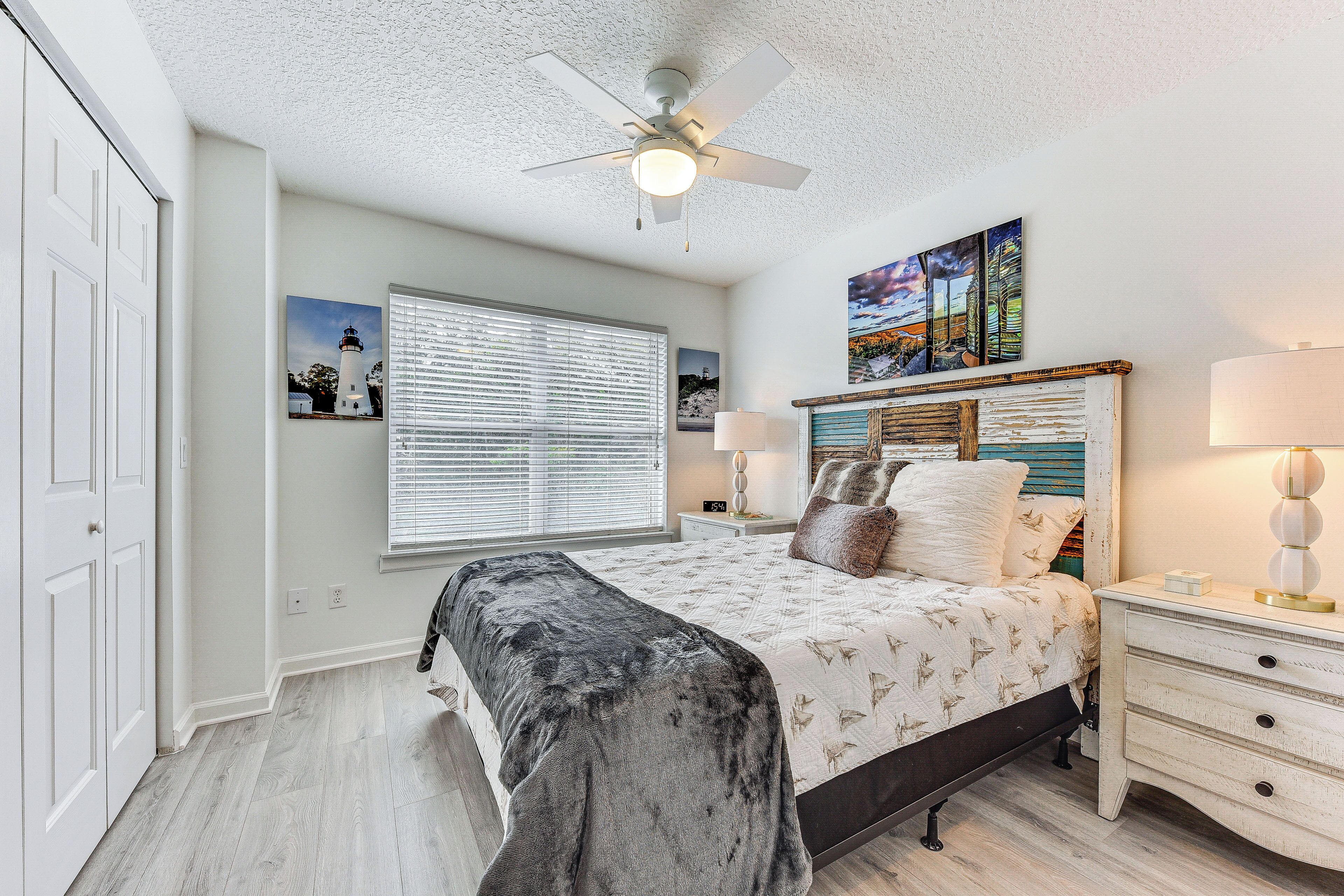A bedroom with a bed, nightstands, a ceiling fan, a window with blinds, and artwork on the wall.