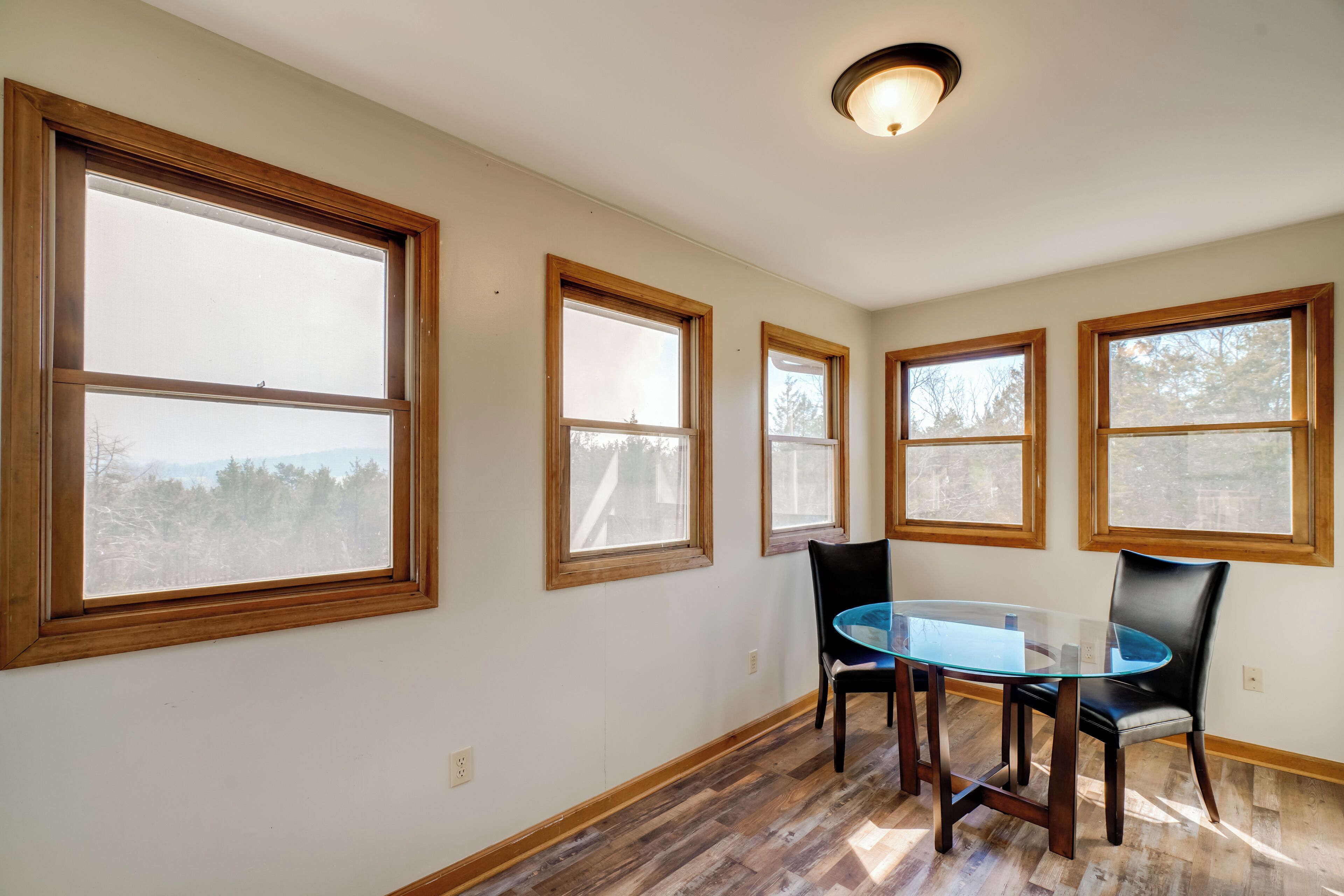 A dining area with a glass table and chairs, large windows with wooden frames, and a ceiling light fixture.