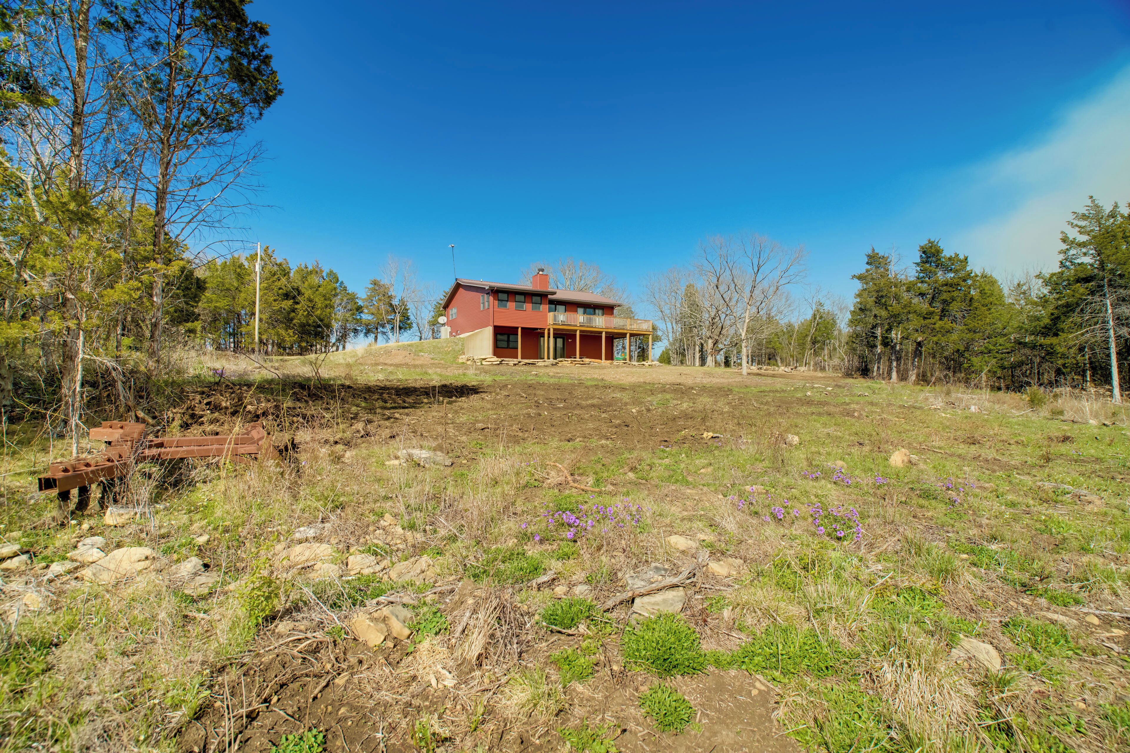 A modern house with a red exterior, a covered porch, and a view of a grassy field with scattered purple flowers.