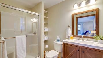 A modern bathroom with a glass-enclosed shower, a toilet, a sink with a mirror, and shelves with towels.