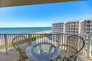A balcony with wicker chairs and a glass table overlooking a beach and a hotel building. - Fort Myers Beach Studio - Sunset & Ocean Views! (Fort Myers Beach)