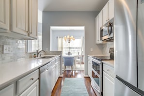 A modern kitchen with white cabinetry, stainless steel appliances, and a blue rug leading to a dining area with a chandelier. - Community Pool & Private Balcony: Chic Lewes Gem (Rehoboth Beach)