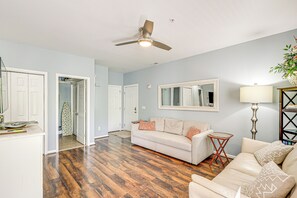 A living room with hardwood flooring, a ceiling fan, a white sofa with patterned cushions, a side table, and a floor lamp. - Community Pool & Private Balcony: Chic Lewes Gem (Rehoboth Beach)