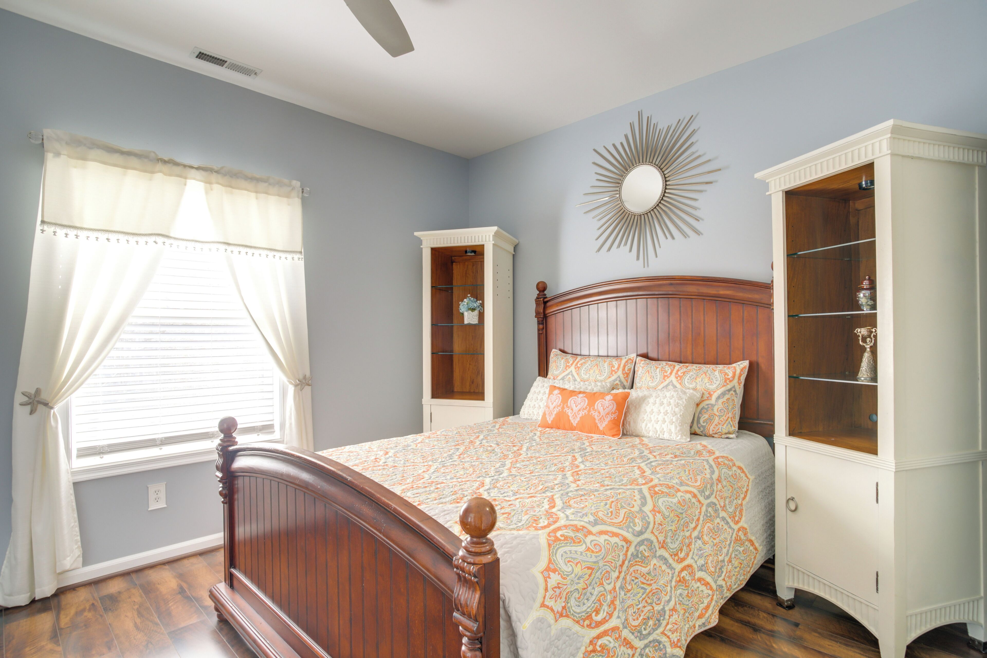 A bedroom with a wooden bed, a window with curtains, a built-in wardrobe, and a wall-mounted mirror.