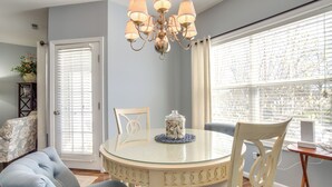A dining area with a round glass table, white chairs, a chandelier, and a view through a window with blinds. - Community Pool & Private Balcony: Chic Lewes Gem (Rehoboth Beach)