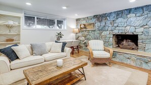 Uma sala de estar com lareira de pedra, sofá branco, poltrona de vime e mesa de centro de madeira.