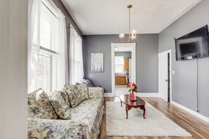 Un salon avec un canapé à motifs floraux, un tapis blanc, une table basse en bois sur laquelle est posé un vase, un téléviseur à écran plat fixé au mur, et une cuisine en arrière-plan.