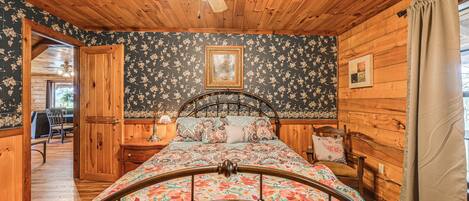 Quarto de cabana de madeira com cama, paredes de madeira, ventilador de teto e porta que dá para outro espaço.
