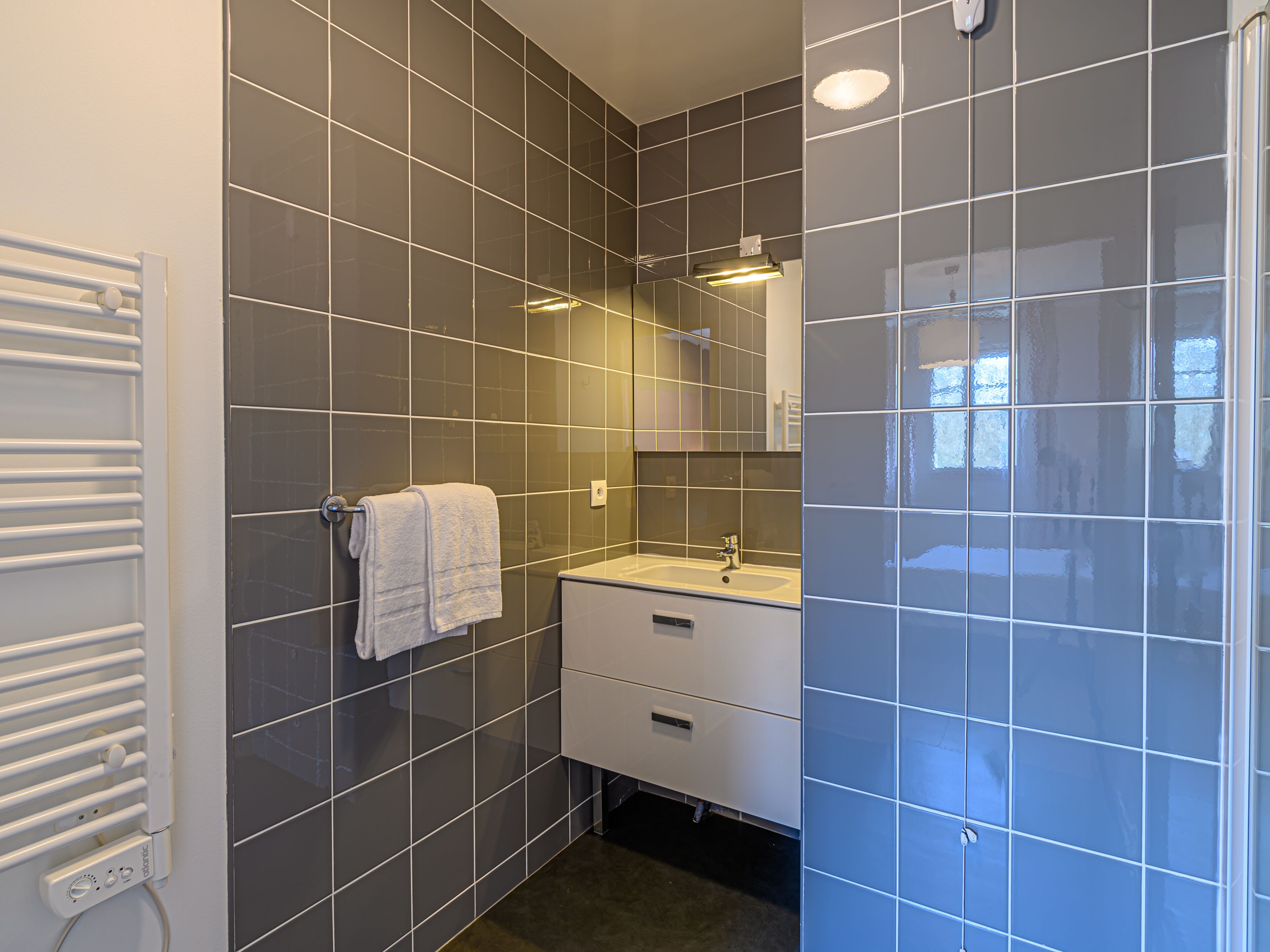 A modern bathroom with grey tiles, a white towel hanging on a rack, a white sink, and a mirror above it.