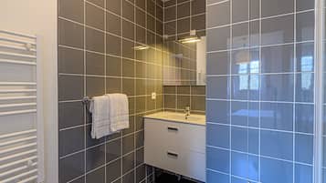 Un baño moderno con baldosas grises, una toalla blanca colgada en un perchero, un lavamanos blanco y un espejo encima.