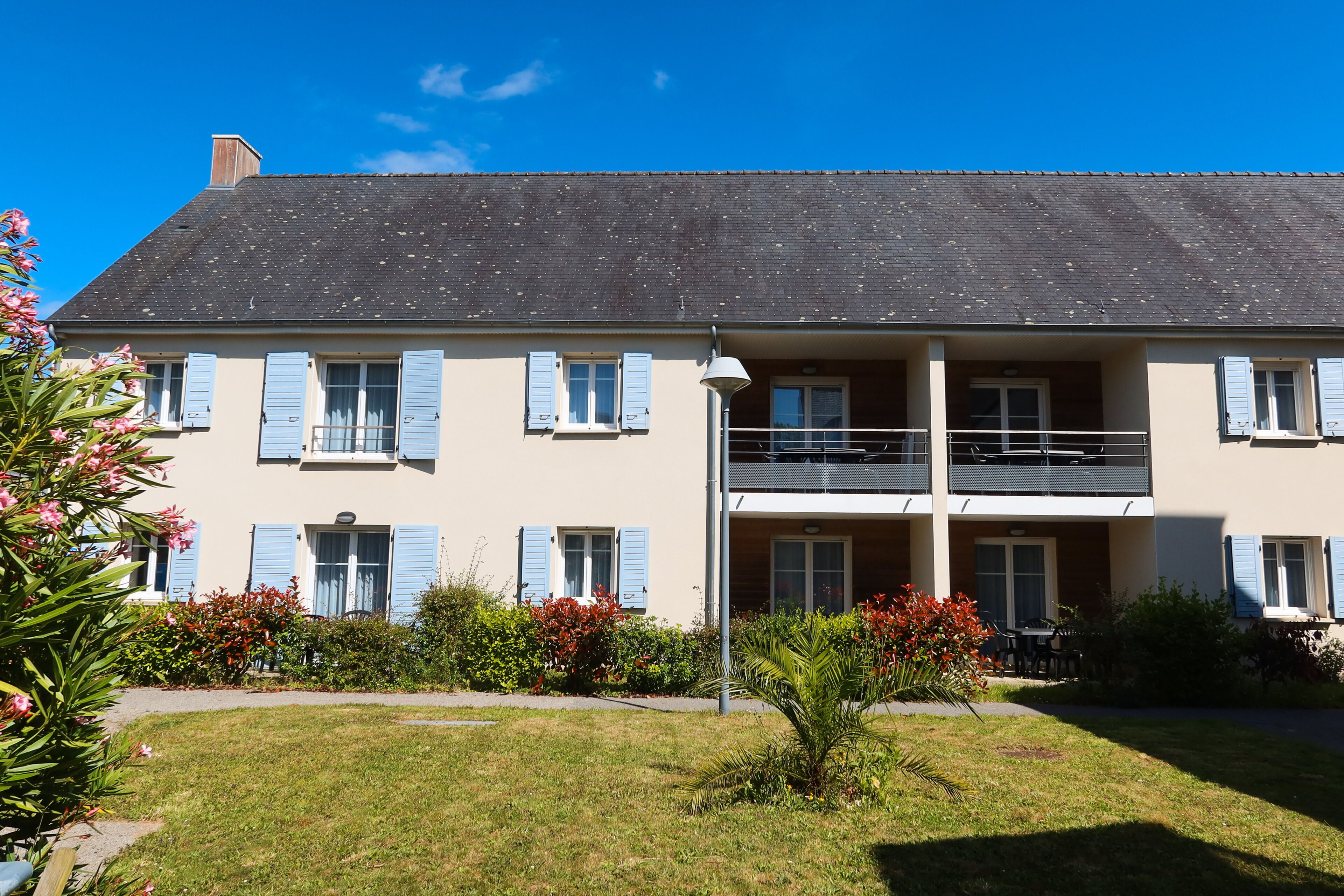 A two-story building with a balcony on the second floor, surrounded by a well-maintained lawn and flowering plants.