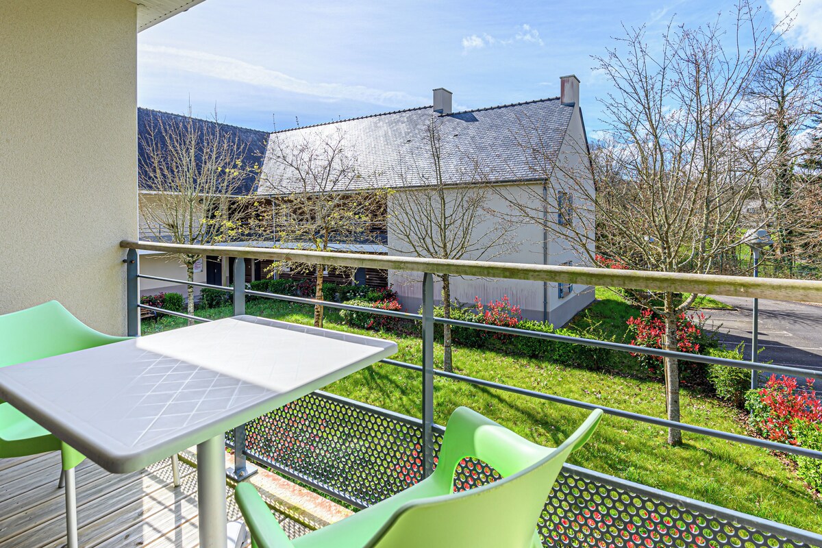 A balcony with a table and chairs, a view of a building with a tiled roof, and a garden with shrubs and flowers.
