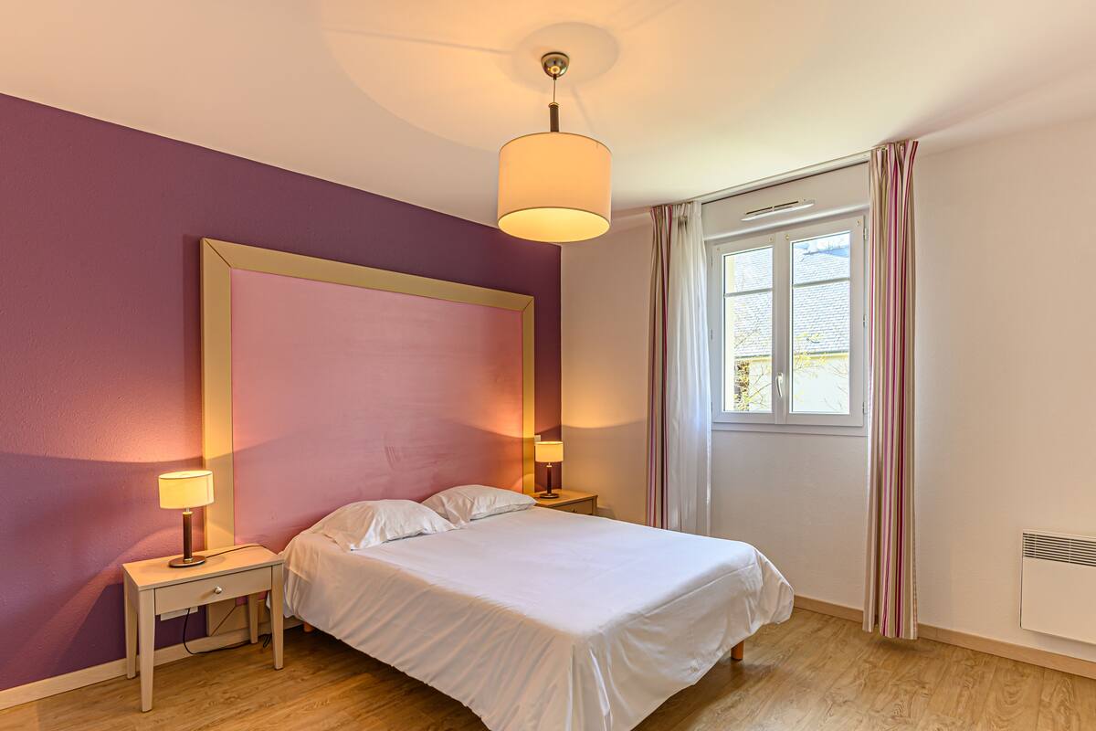 Ein Schlafzimmer mit einem Bett, Nachttischlampen, einem Fenster mit Vorhängen und einer Deckenleuchte.