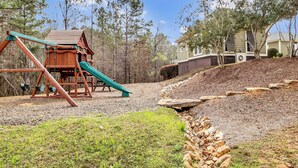 A playground with a swing set and slide, surrounded by trees and a house in the background. - Balcony + Community Perks: Lake Martin Condo! (Dadeville)