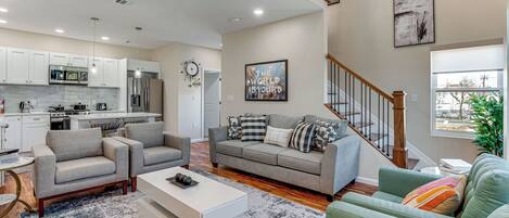 A modern living room with a grey sofa, a white coffee table, and a green sofa. There is a kitchen in the background with white cabinets and stainless steel appliances.