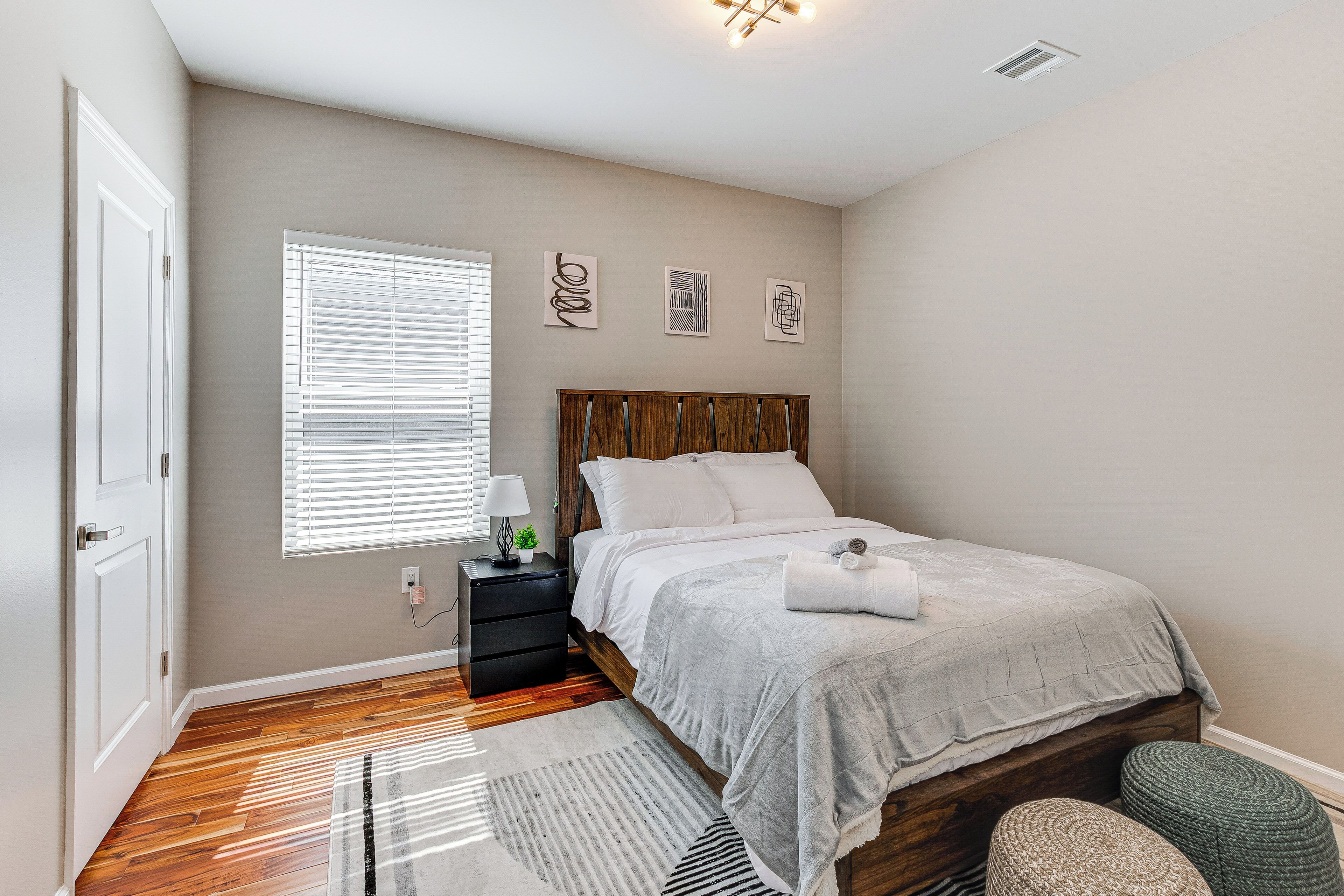 A bedroom with a wooden headboard, a bed with white bedding, a nightstand with a lamp, and a window with blinds.
