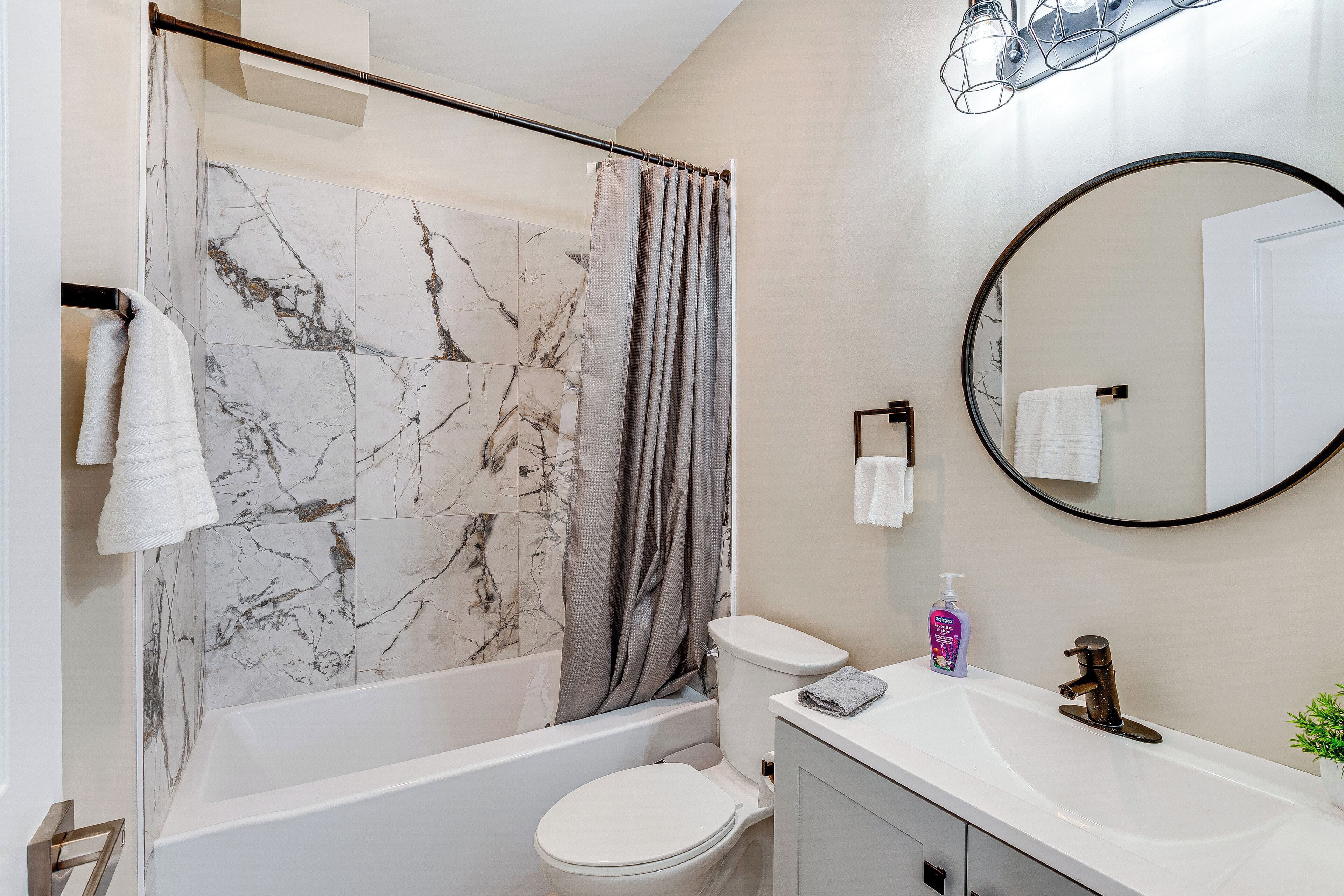 Une salle de bain moderne avec un mur de douche en marbre, une baignoire blanche, des toilettes, un lavabo avec un miroir et une petite plante en pot.