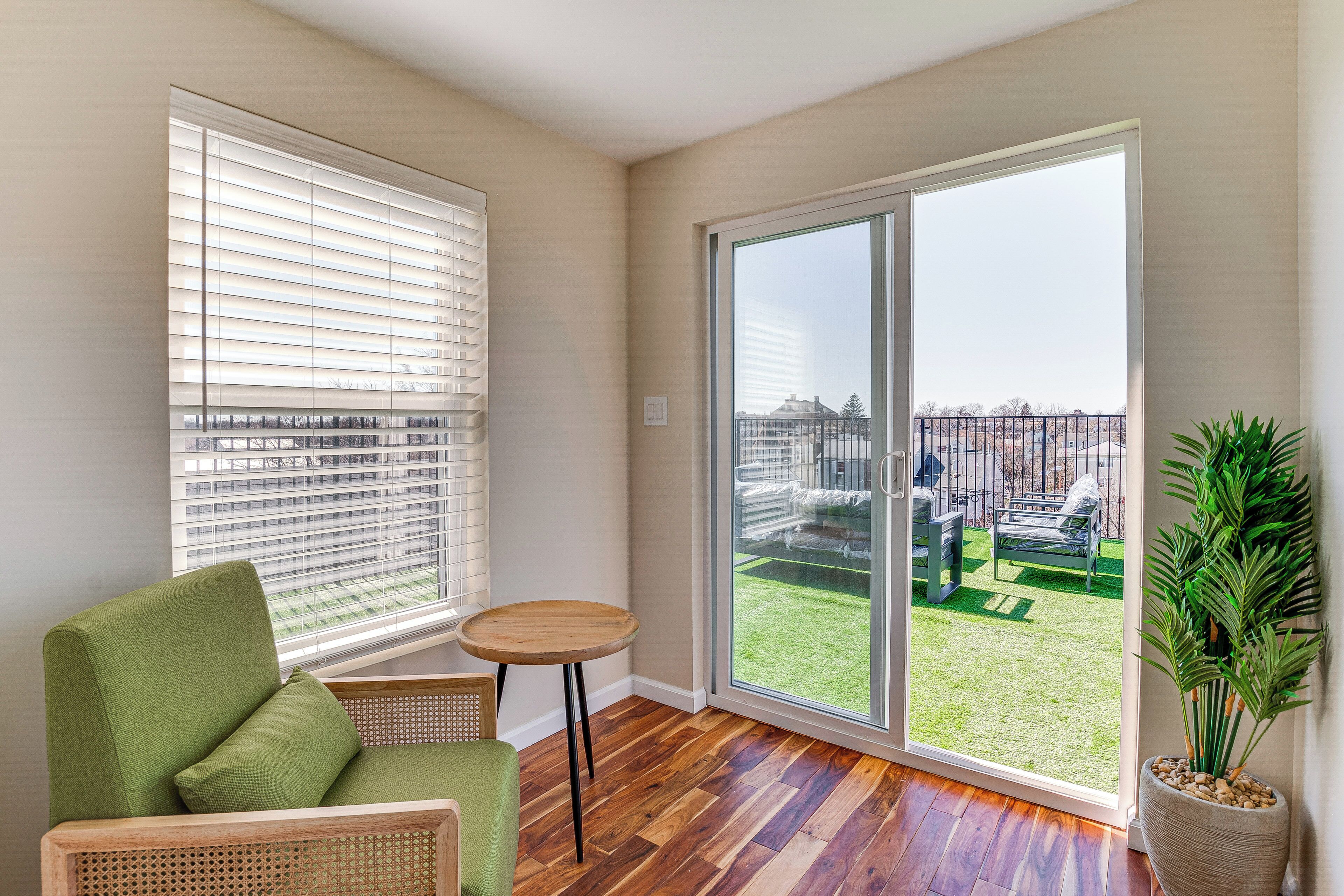 Een kamer met een groene bank, een klein houten tafeltje en een grote schuifglazen deur die uitkomt op een balkon met uitzicht op een grasveld en gebouwen.